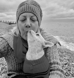 Close-up of mature woman kissing dog at beach against cloudy sky