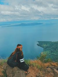 Side view of young woman with backpack sitting on mountain by sea against cloudy sky