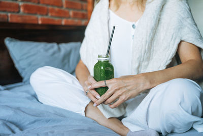 Crop photo of young woman in casual clothes with green smoothie sitting on bed at home
