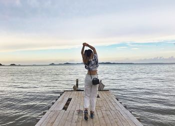 Rear view of woman with arms raised standing on pier in lake against cloudy sky