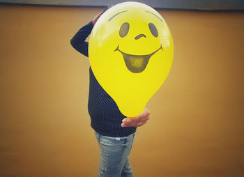 Midsection of man holding yellow balloon with anthropomorphic face against wall