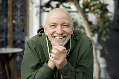 Senior man with beard joyfully clasping hands in green jacket outdoors