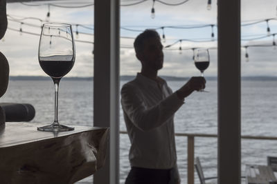 Man holding drink in glass while standing by sea against sky