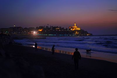 Silhouette people on israeli beach by illuminated buildings against sky during sunset in tel-aviv