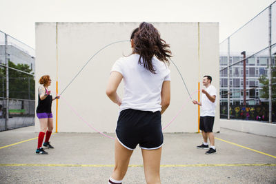 Woman walking towards friends holding jump ropes on street against wall