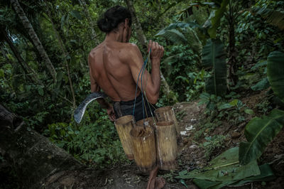 Rear view of shirtless man carrying wooden containers while walking amidst trees in forest