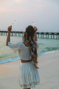 Rear view of woman with arms raised standing at beach against sky