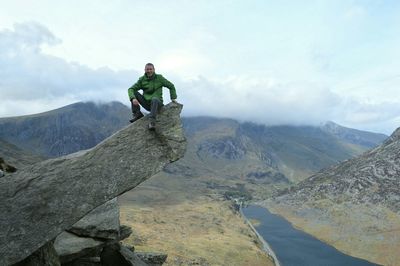 Full length of mature man sitting on cliff against mountain range