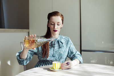 Beautiful elegant woman sits in the kitchen and pouring wine from bottle to the teacup