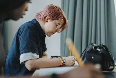 Side view of young non-binary person writing while sitting at desk in classroom