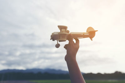 Cropped hand of person holding model airplane against cloudy sky