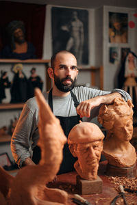 Pensive bearded male woodworker in apron standing at table with carved wooden sculptures and professional instruments during work in joinery