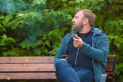 Man smoking electronic cigarette while sitting on bench against tree