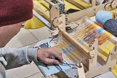 Hand of older man, who weaving small rug with pattern on wooden manual table loom.