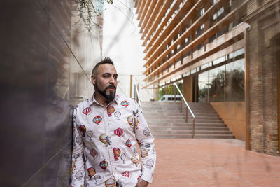 Bearded male with hand on pocket leaning on wall, looking camera