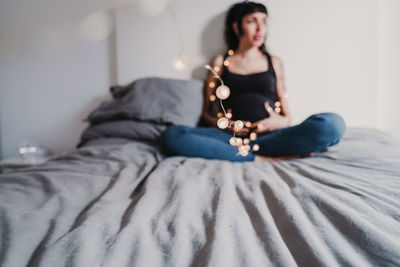 Pregnant young woman sitting with string lights on bed at home