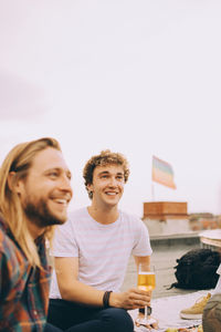 Smiling male friends enjoying beer while looking away at terrace during party against sky