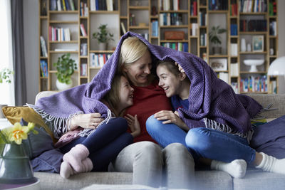 Mother and her daughters cuddling and having fun, sitting on couch, covered in blanket