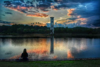 Woman sitting by lake against sky during sunset
