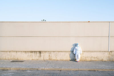 Nurse wearing protective suit standing on footpath against wall