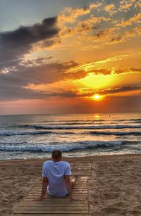 Rear view of man sitting at beach against cloudy sky during sunset