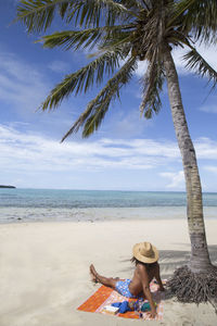 Man with sunhat and curly hair chilling under palm tree at sandy beach
