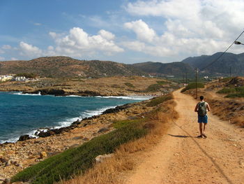 Rear view of man walking on dirt road by sea against sky