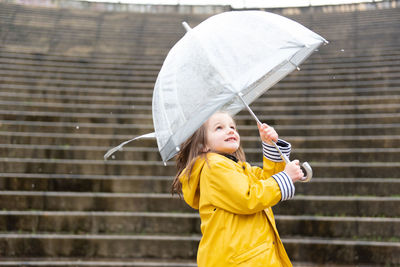 Playful kid in yellow raincoat and with wet umbrella standing on street enjoying rainy weather in city