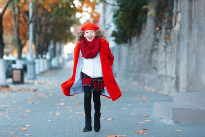 Happy kid girl 5-6 year old with blonde hair wear red jacket and beret hat walk in city street.