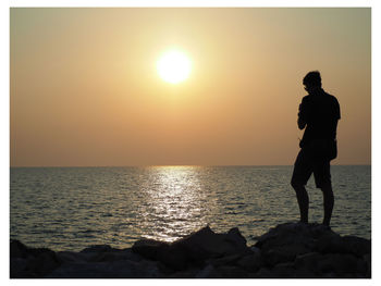 Silhouette man standing in sea against sky during sunset