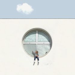 Girl sitting on window against sky