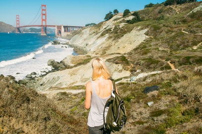 Rear view of woman standing at sea shore by golden gate bridge against clear blue sky