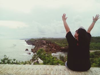 Rear view of carefree woman with arms raised sitting on retaining wall with sea in background