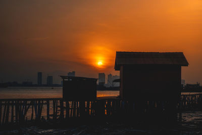 Silhouette buildings by sea against orange sky