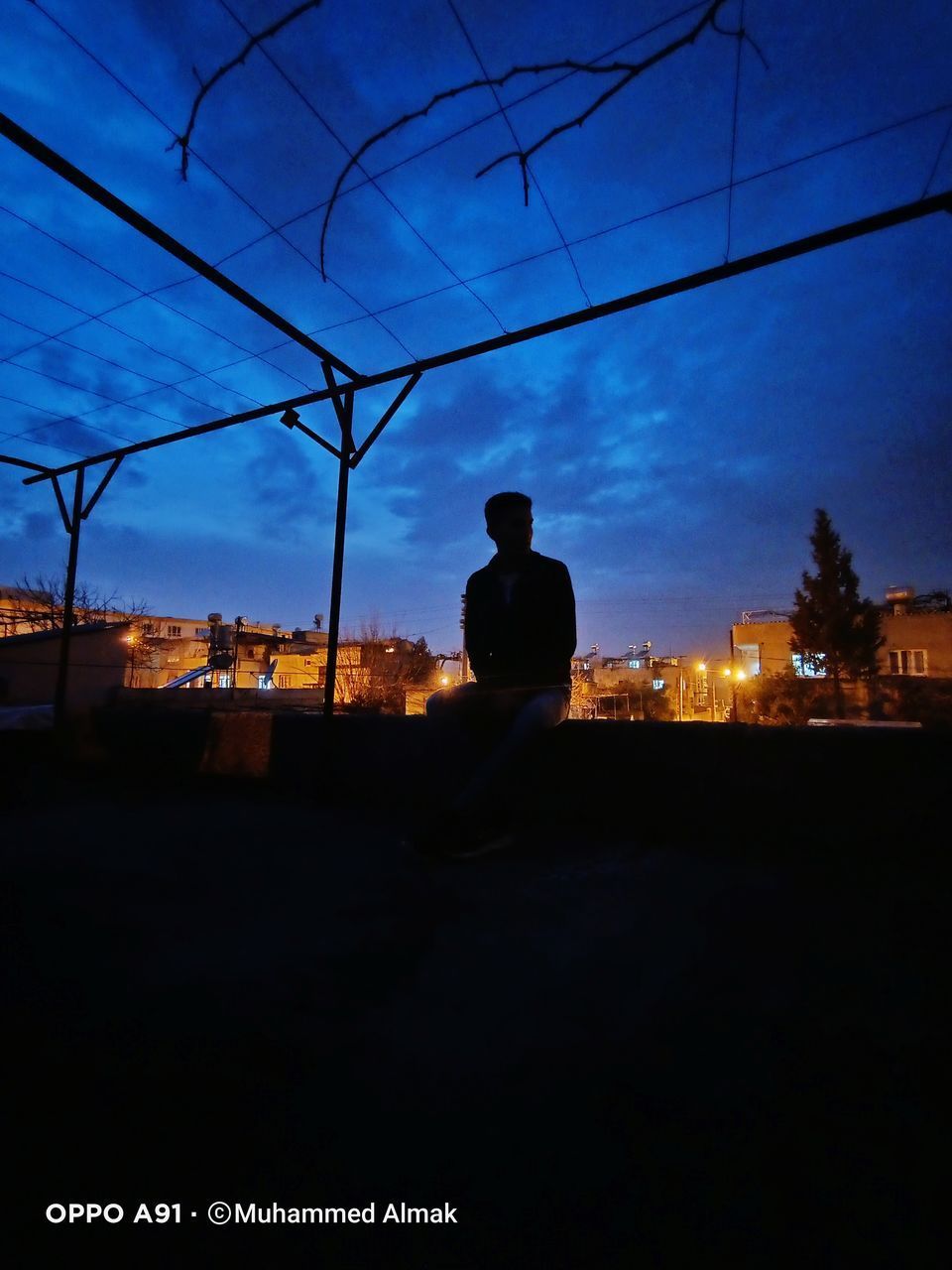 SILHOUETTE MAN SITTING ON CITY AGAINST SKY AT SUNSET