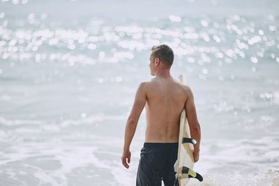 Rear view of shirtless man with surfboard standing at beach