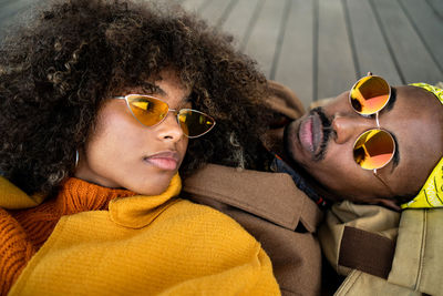 Top view of trendy african american man and woman in bright coat lying down on wooden floor looking at each other