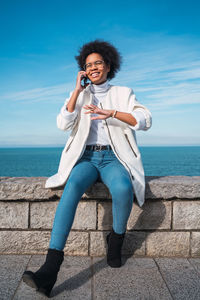 Smiling young woman talking over mobile phone on retaining wall against sea