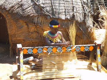 Woman weaving straw while sitting against hut