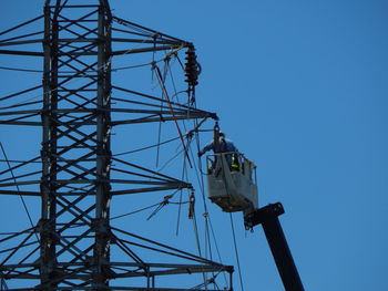 Worker on cherry picker repairing electricity pylon against clear blue sky