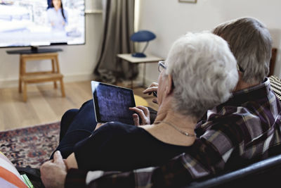 Rear view of senior couple sitting with digital tablet watching tv in living room at home