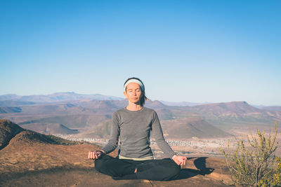 Woman practicing yoga on rock at mountain peak against blue sky during sunny day