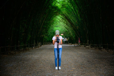 Woman using camera while standing on road against trees
