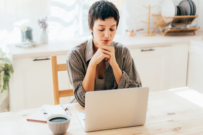 Woman freelancer sitting in the kitchen at home working on a laptop.