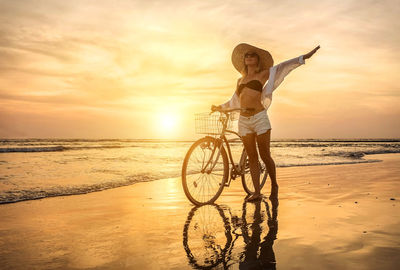 Side view of woman riding bicycle on beach against sky during sunset