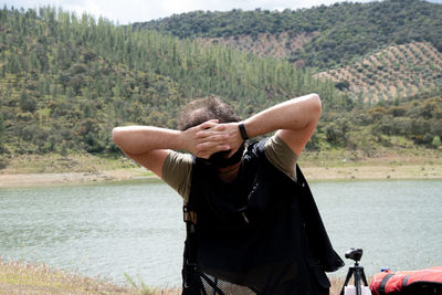 Full length of man with hands behind head sitting at lakeshore against mountains