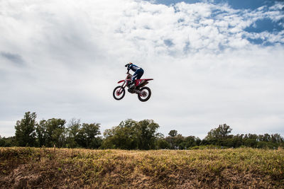 Low angle view of man jumping motorcycle over field against cloudy sky