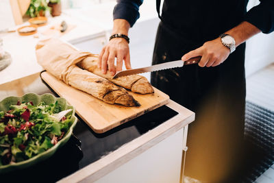 Midsection of man cutting bread by salad at kitchen counter