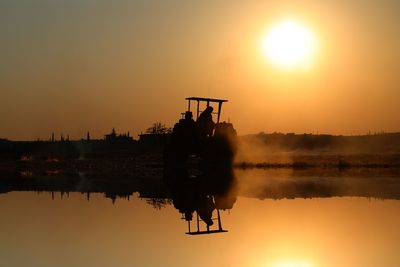 Silhouette woman sitting on tractor by lake against sun in orange sky