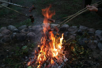 Cropped hand cooking sausages on campfire at dusk